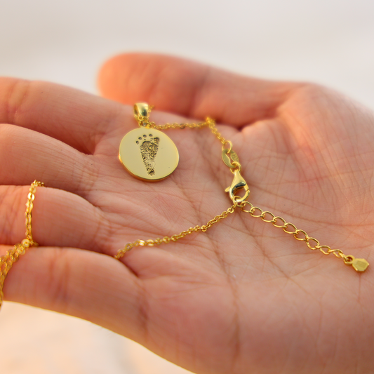 Gold Aurelia round necklace engraved with a baby footprint, displayed in the palm of a hand against a soft neutral background.