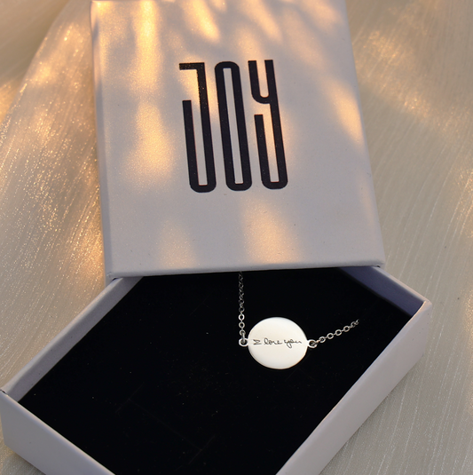 Personalized silver handwriting bracelet with round engraved charm displayed inside an open Chapters of Joy jewelry box on soft ivory fabric.