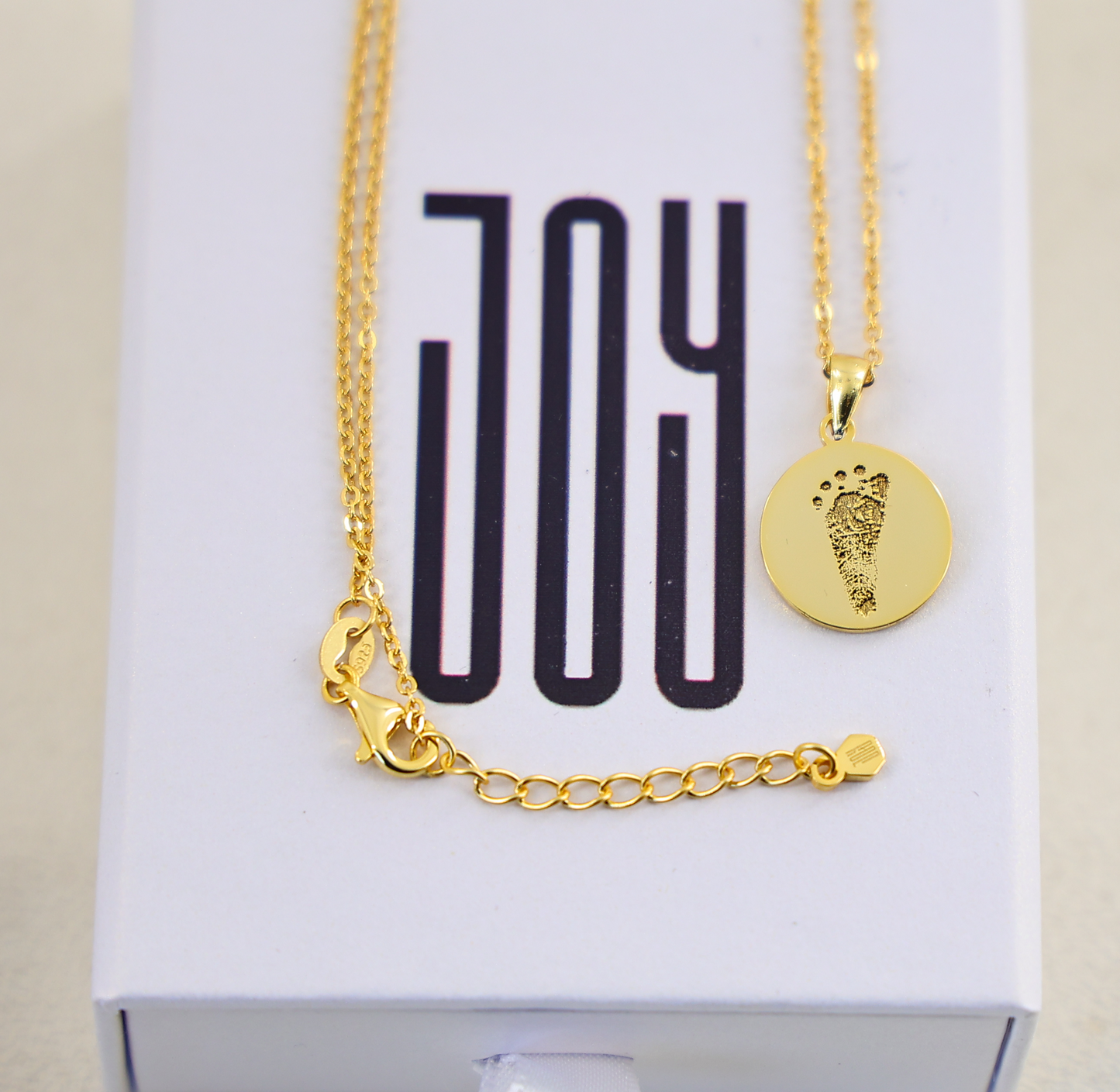 Gold necklace with a round pendant on a white box with 'JOY' printed on it.