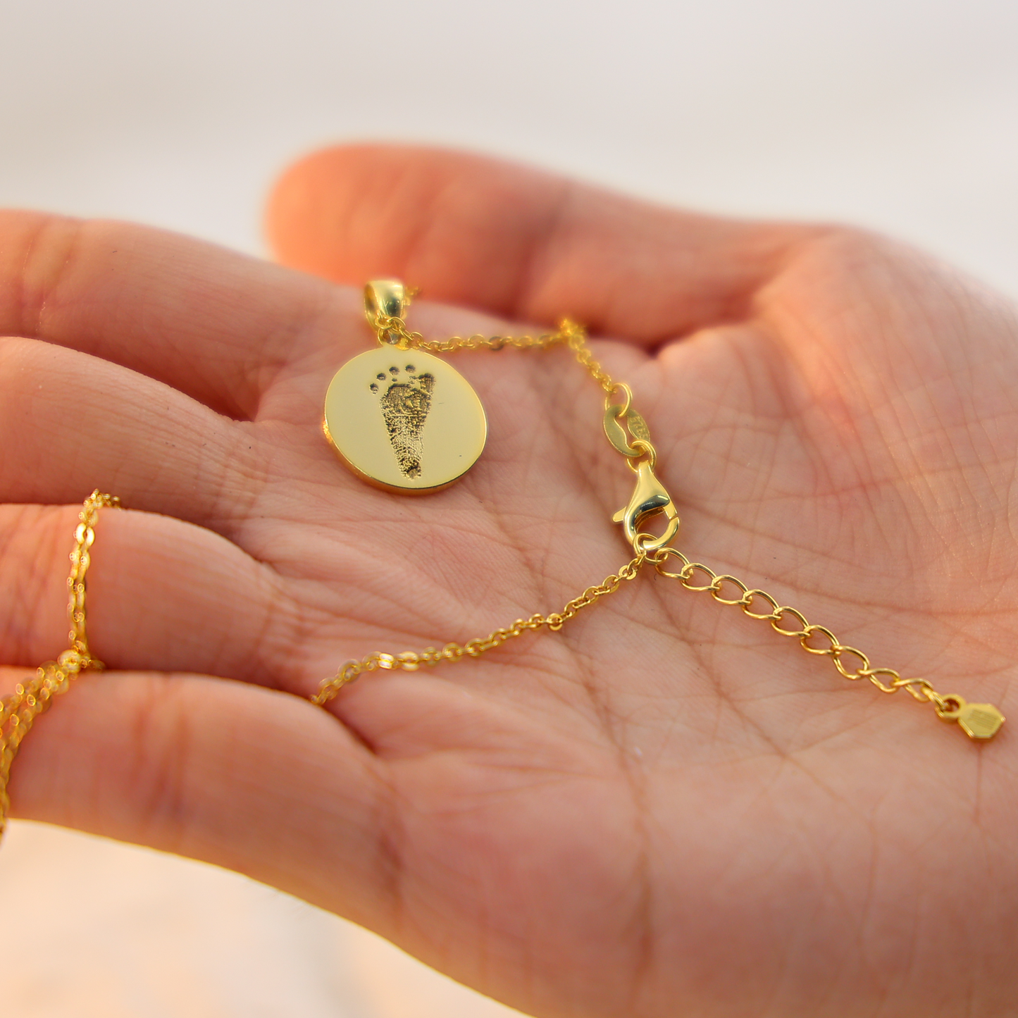 Gold Aurelia round necklace engraved with a baby footprint, displayed in the palm of a hand against a soft neutral background.