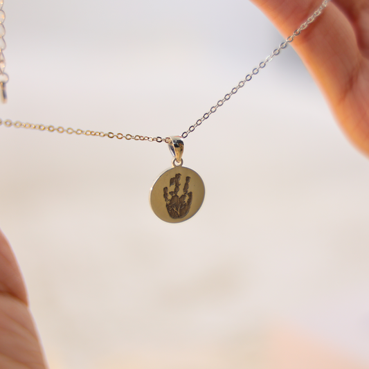 Round sterling silver Aurelia necklace engraved with a handprint, suspended on a fine chain and held between two hands against a soft neutral background.